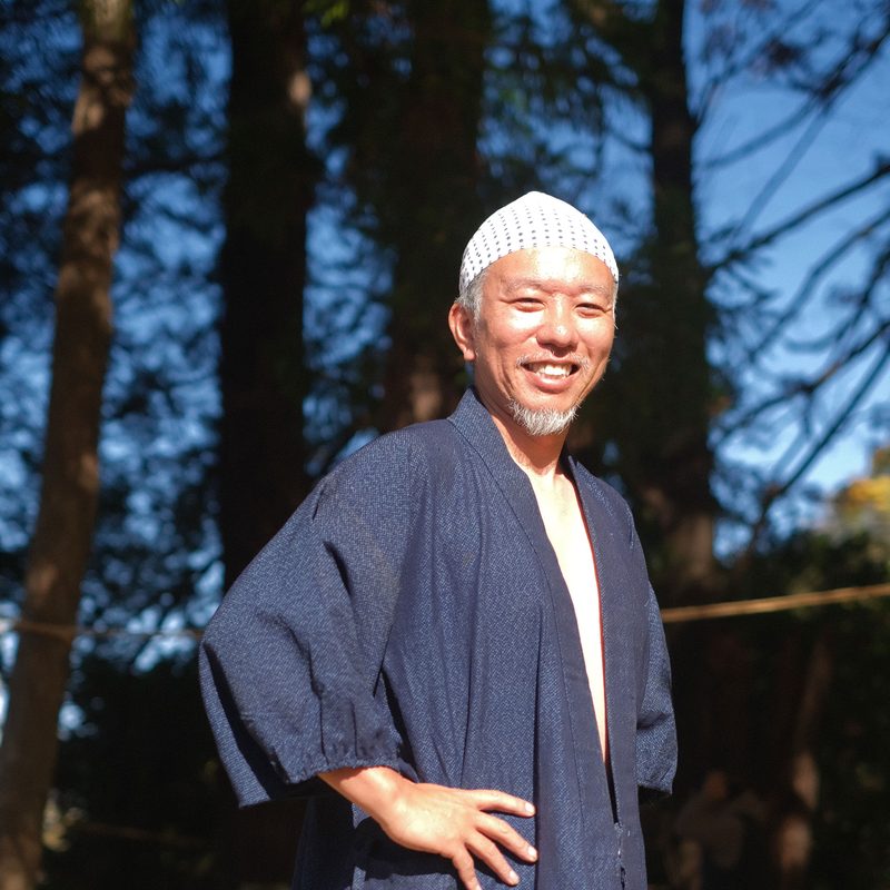 古谷如弘。紺の作務衣に白い帽子で、木立の前に立って笑っている
